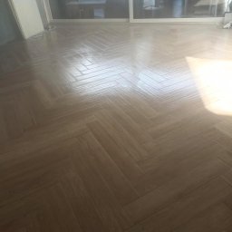 Tanio i z fakturką - Ułożona w jodełkę, błyszcząca, brązowa podłoga z płytek imitujących drewno, świeżo po położeniu, z widocznymi refleksami światła.