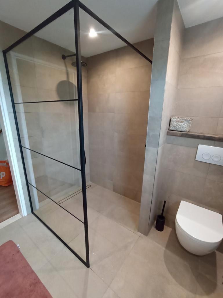 Nowoczesna kabina prysznicowa z czarnymi profilami, odpływ liniowy w płytkach, beżowe płytki ścienne i podłogowe, wisząca toaleta, minimalistyczny design.