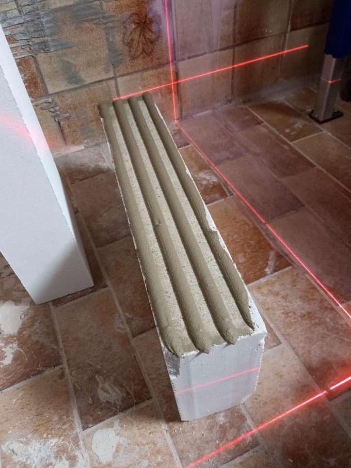 Bloczek z betonu komórkowego z naniesioną zaprawą klejową, widoczne linie lasera poziomującego, przygotowany do montażu na podłodze wyłożonej płytkami imitującymi drewno.