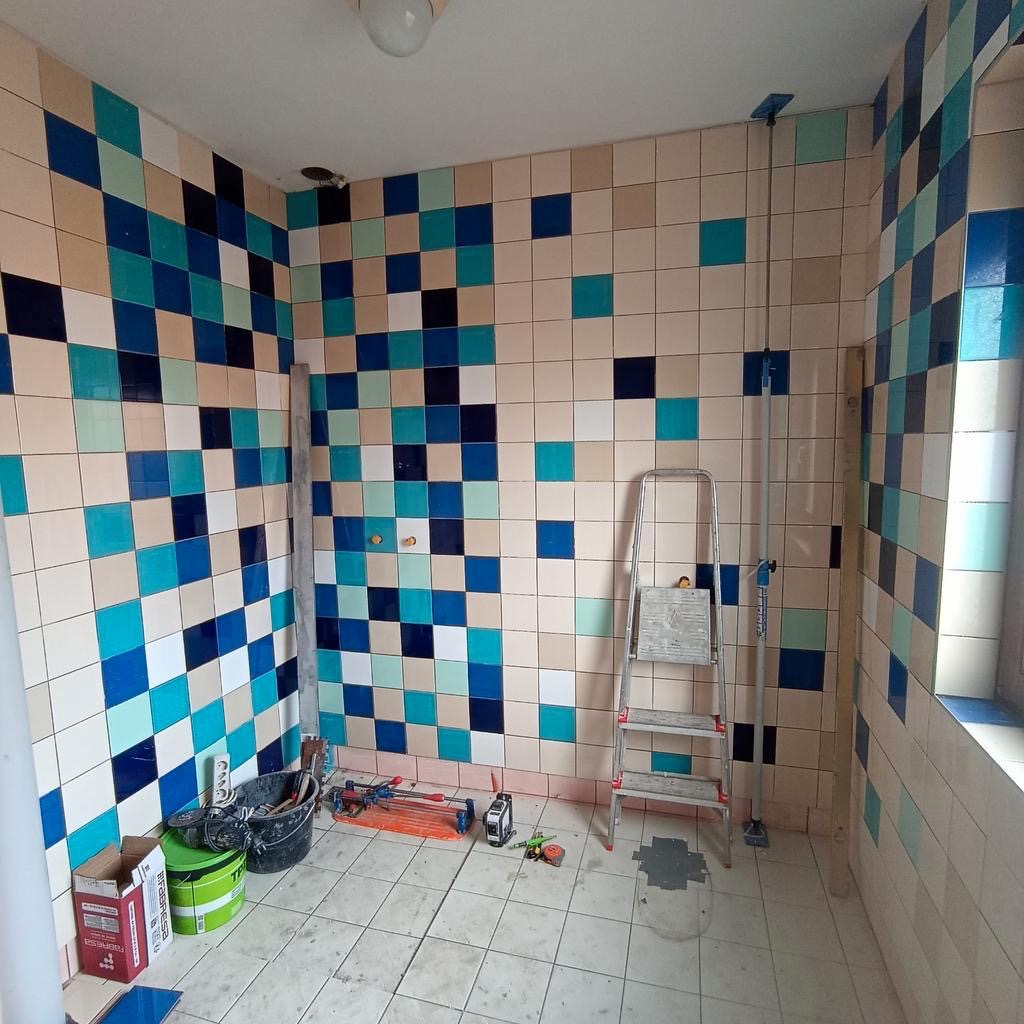 Niedokończona łazienka z mozaiką z kwadratowych płytek w odcieniach błękitu, beżu i granatu, widoczna drabina, narzędzia glazurnicze i opakowania po kleju do płytek.