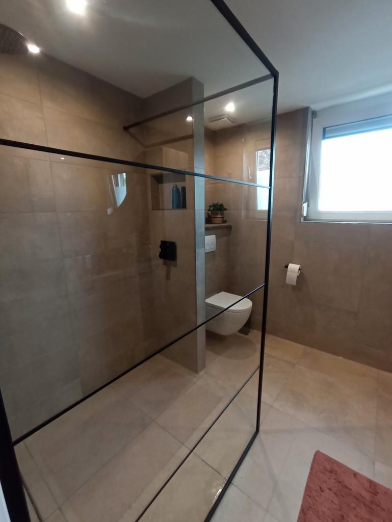 Nowoczesna łazienka z kabiną prysznicową typu walk-in z czarnymi profilami, beżowe płytki na ścianach i podłodze, wisząca toaleta, półka z dekoracjami.