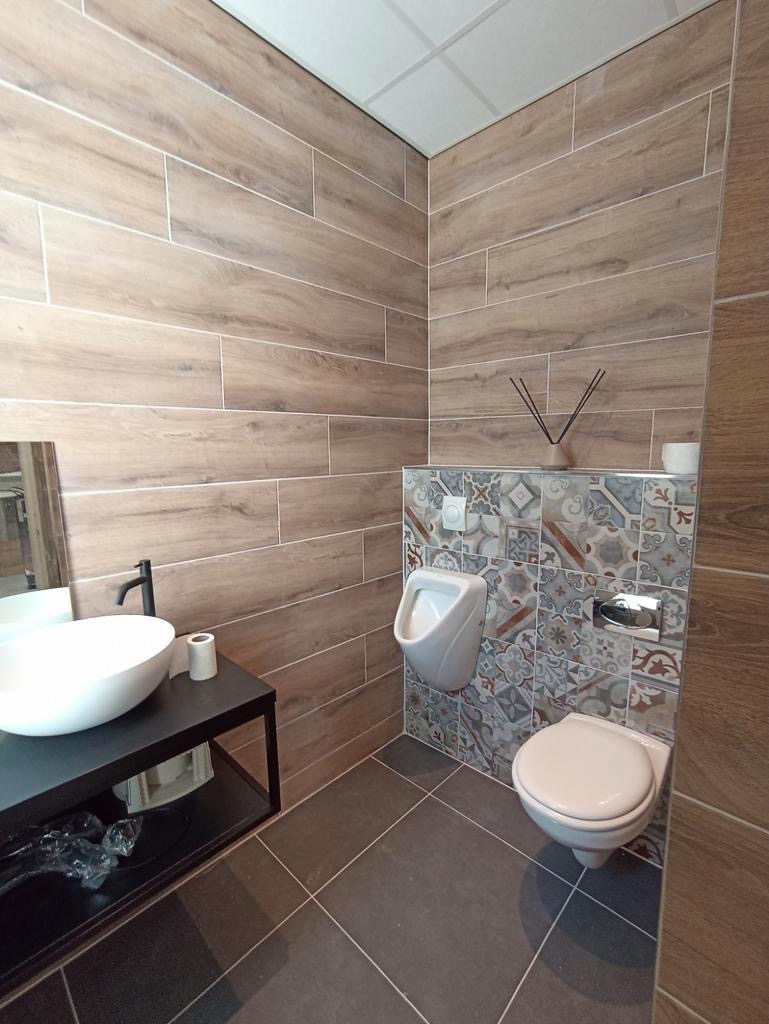 Wykończona łazienka z drewnopodobnymi płytkami ściennymi, wiszącą miską WC i pisuarą na ścianie z dekoracyjnymi płytkami, ciemnoszara podłoga z płytek, biała umywalka nablatowa na czarnym stelażu.