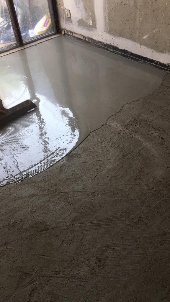 Wyrównywanie podłogi w pomieszczeniu za pomocą szpachli, widoczny etap wylewania wylewki samopoziomującej, w tle okno i fragment ściany z widocznymi śladami po wcześniejszych pracach.