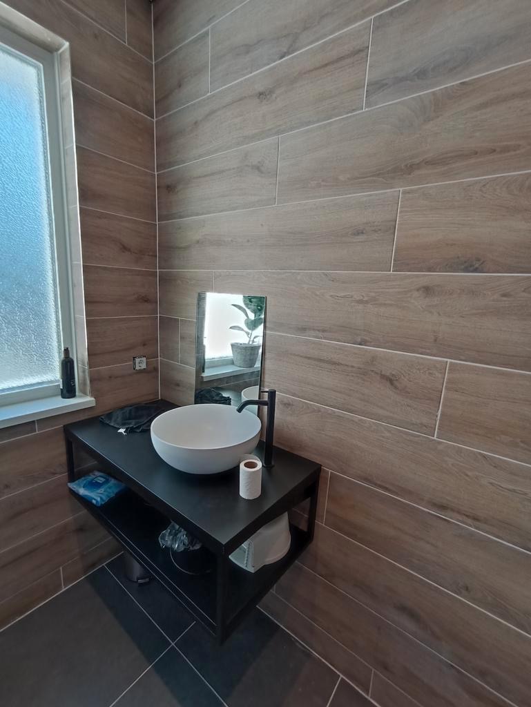 Nowoczesna łazienka z drewnopodobnymi płytkami na ścianach, okrągłą białą umywalką nablatową na czarnym metalowym regale i czarną armaturą.