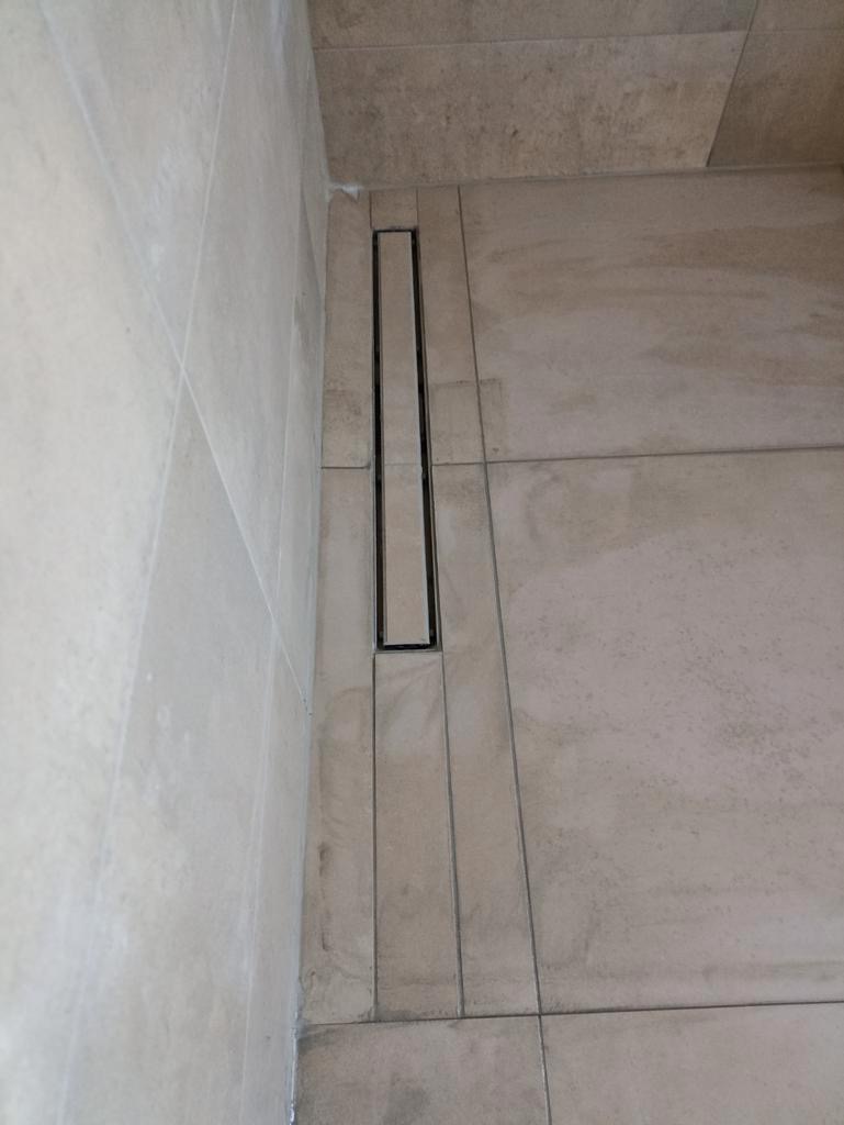 Beżowe płytki podłogowe i ścienne z liniowym odpływem prysznicowym w kabinie typu walk-in, widok z góry.