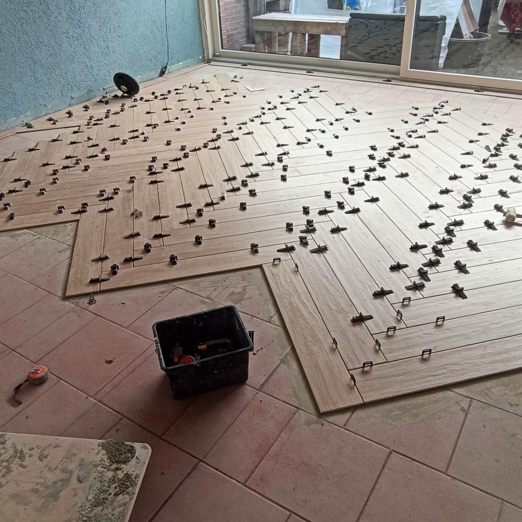 Układanie podłogi z płytek imitujących drewno w jodełkę, z użyciem klipsów poziomujących, widoczny fragment starej podłogi i pojemnik z narzędziami.