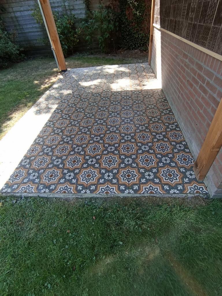 Wyłożony wzorzystymi płytkami taras przylegający do ściany budynku, z widoczną konstrukcją z drewna i fragmentem trawnika.