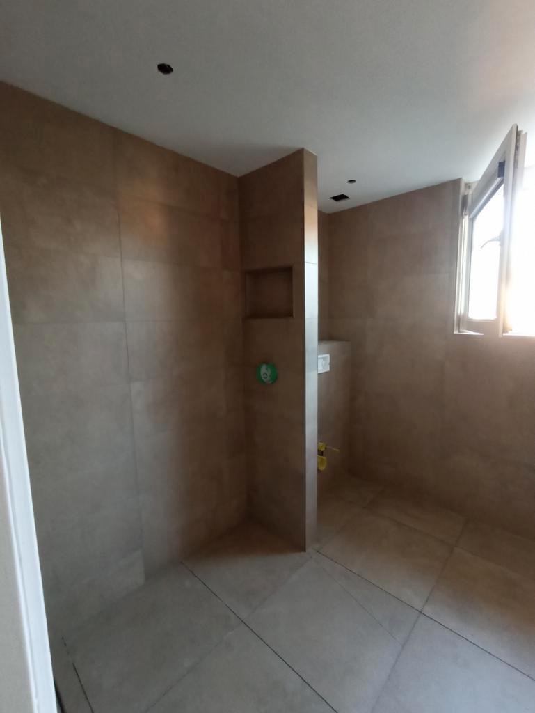 Beżowa, wykafelkowana łazienka z wnęką w ścianie, otwartym oknem i widocznymi instalacjami hydraulicznymi przed montażem armatury.