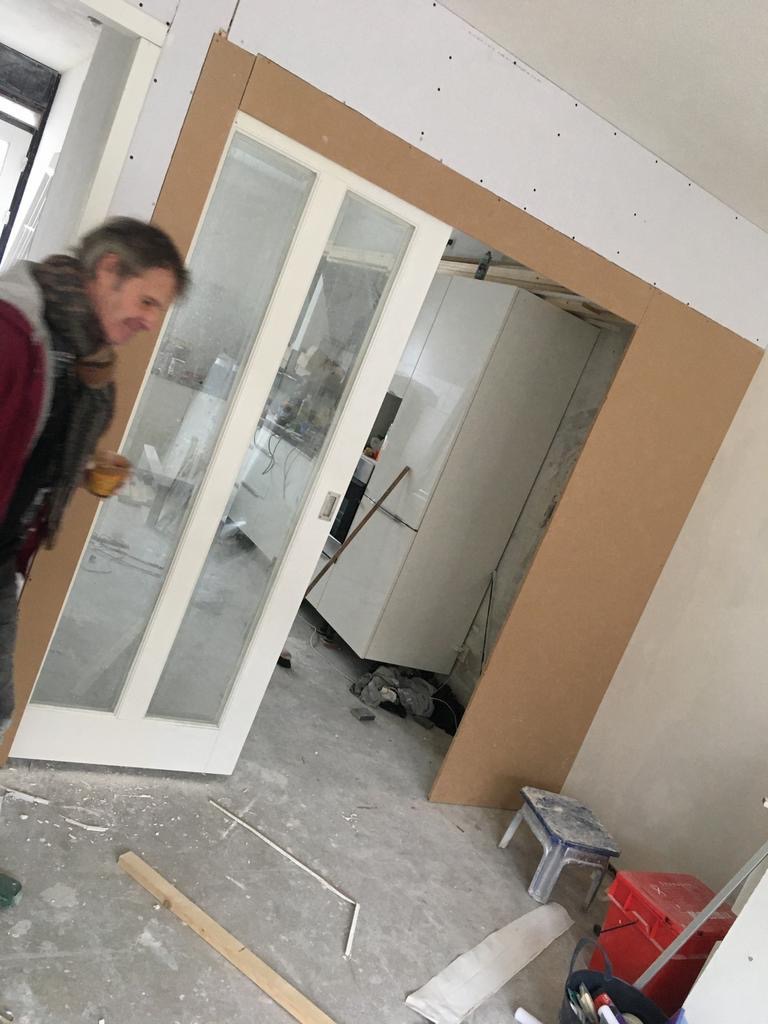 Montaż szklanych drzwi przesuwnych w ościeżnicy z płyty karton-gips, widoczny fragment kuchni w tle oraz narzędzia i materiały budowlane na podłodze.