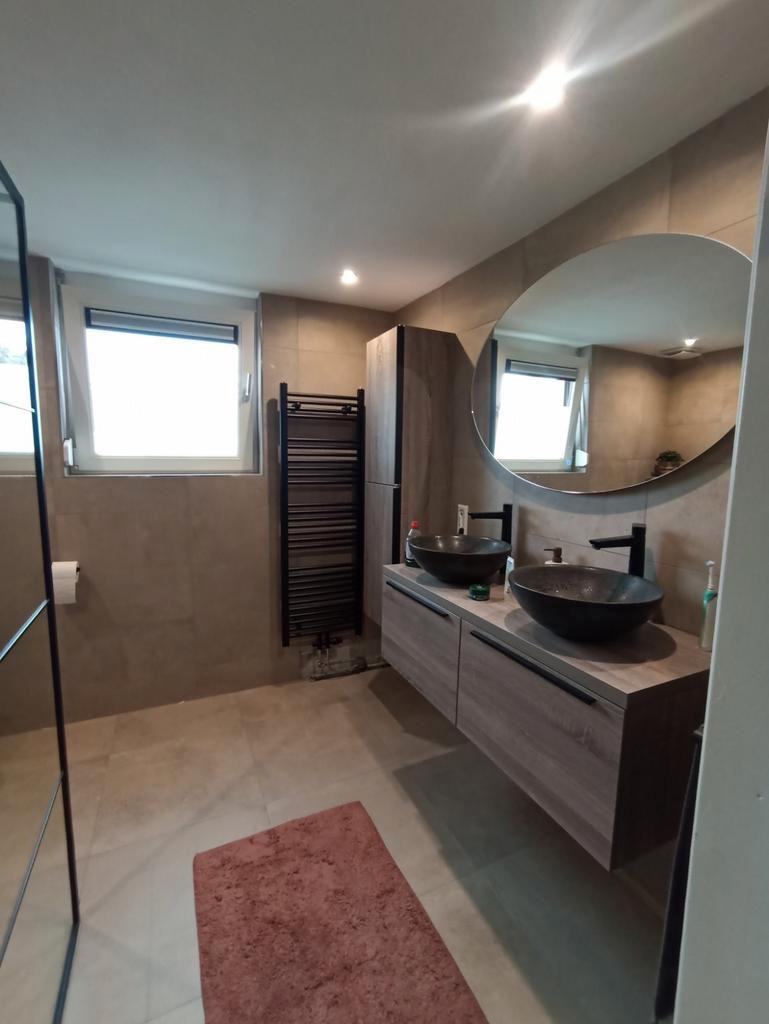 Nowoczesna łazienka z dwoma okrągłymi umywalkami nablatowymi w kolorze grafitowym, szafką pod umywalką w odcieniach drewna, dużym okrągłym lustrem bez ramy i grzejnikiem drabinkowym w kolorze czarnym.