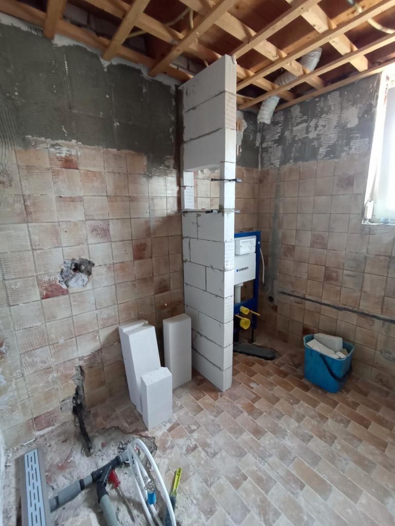 Demontaż starej łazienki z widoczną instalacją podtynkową Geberit, surowymi ścianami i podłogą z ceglastych płytek. Częściowo wzniesiona ścianka działowa z bloczków betonu komórkowego.