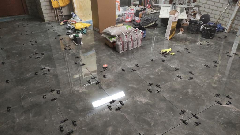 Ułożone, błyszczące, szare płytki podłogowe z krzyżykami dystansowymi, worki z klejem w tle, etap prac wykończeniowych.