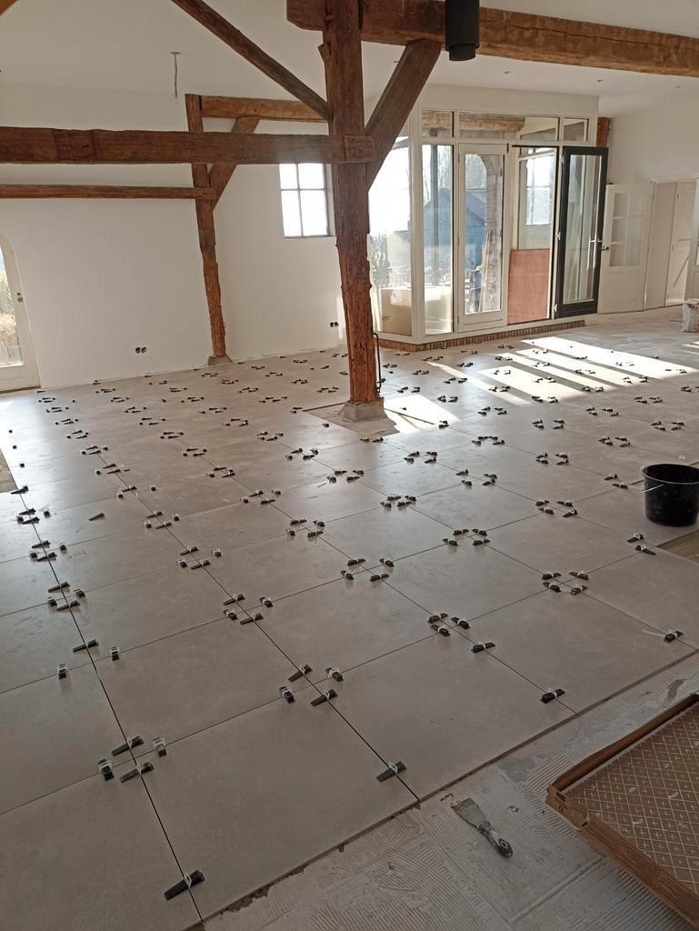 Układanie jasnych, kwadratowych płytek podłogowych w przestronnym, jasnym pomieszczeniu z drewnianymi belkami stropowymi, z użyciem klipsów poziomujących do spoin.