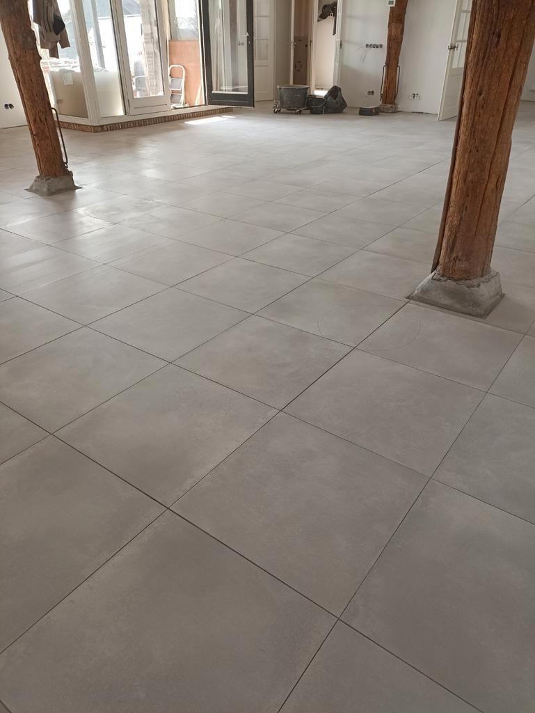 Ułożone szare płytki podłogowe w dużym pomieszczeniu z drewnianymi słupami nośnymi, widoczne drzwi balkonowe i przygotowania do dalszych prac wykończeniowych.