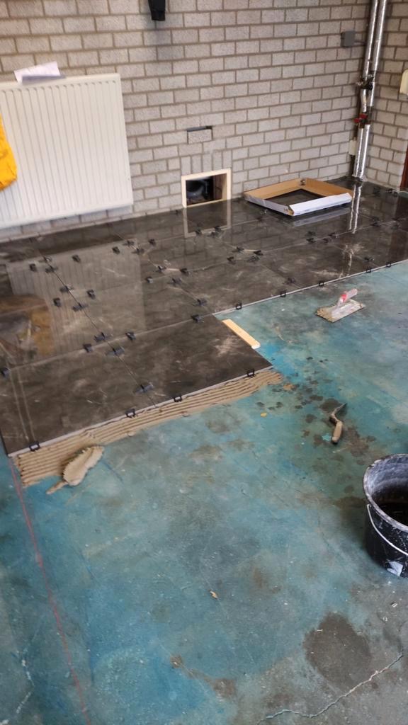 Układanie ciemnych, marmurkowych płytek podłogowych z użyciem klipsów poziomujących, widoczny fragment ceglanej ściany i rury instalacyjne.