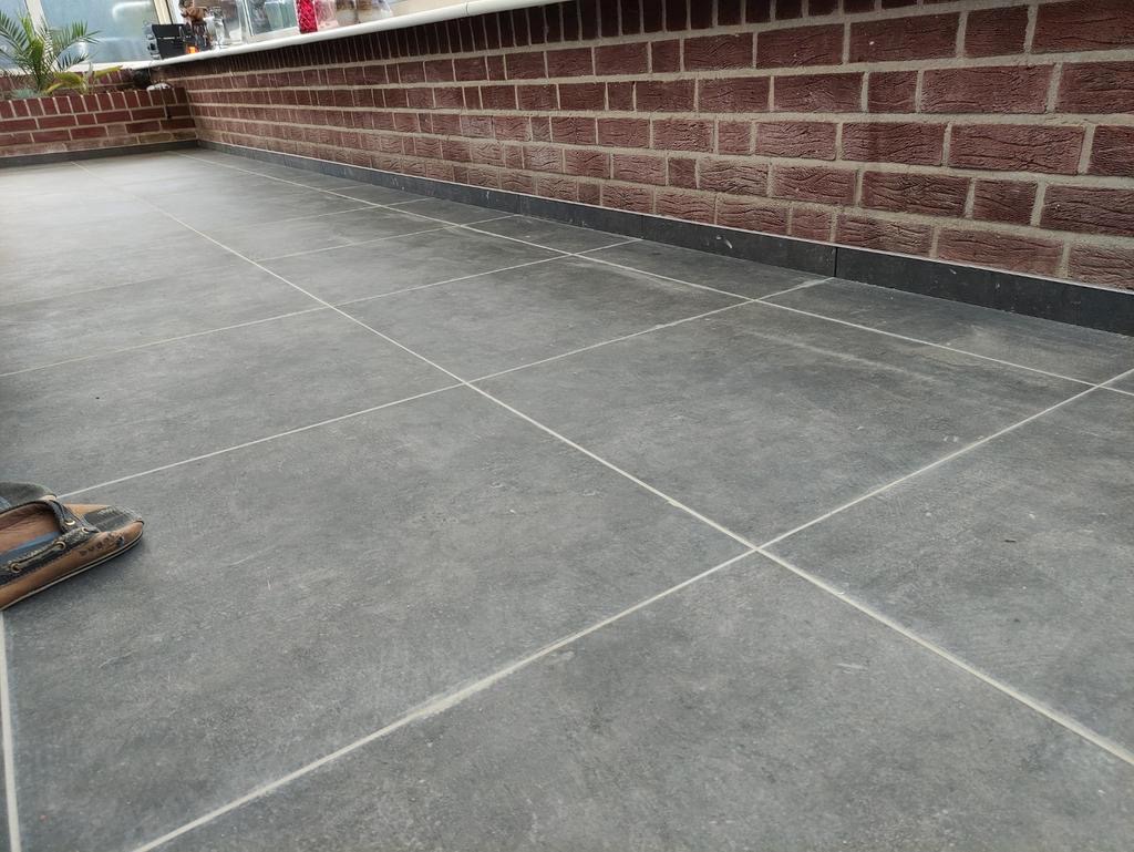 Ułożona szara terakota podłogowa z fugami, przy ceglanej ścianie z dekoracyjnym parapetem, widoczny fragment buta w lewym dolnym rogu.