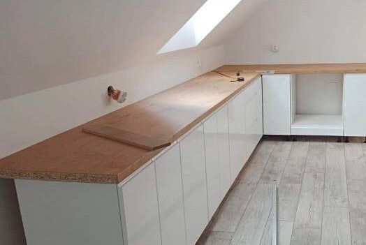 Białe szafki z drewnianym blatem w skosie poddasza, widoczny fragment niezamontowanej deski, narzędzie pomiarowe na blacie.