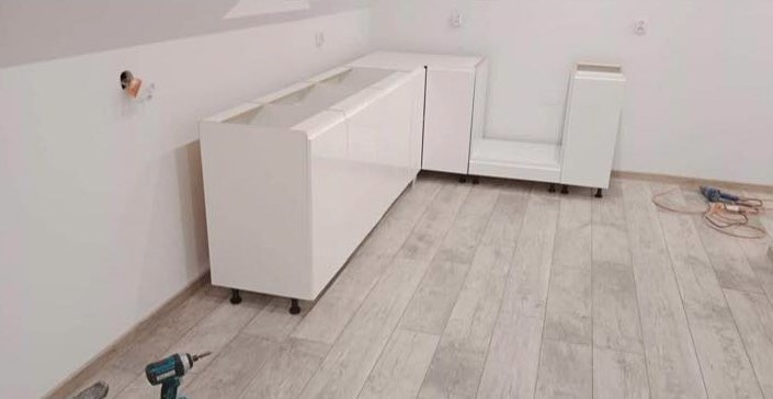 Białe, lakierowane szafki kuchenne bez blatów, ustawione w rogu pomieszczenia na jasnej podłodze z paneli, z widocznymi narzędziami montażowymi w tle.