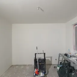 Świeżo pomalowane na biało wnętrze pokoju z zabezpieczonym oknem folią malarską, widoczne narzędzia malarskie na podłodze i w wózku narzędziowym.