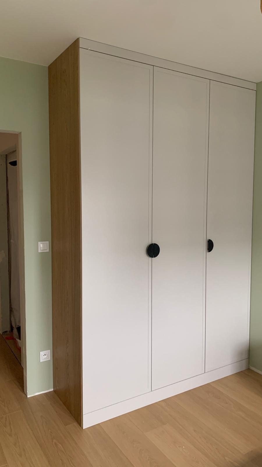 Trzydrzwiowa szafa wnękowa z białymi frontami i okrągłymi, czarnymi uchwytami, z widoczną boczną ścianą z imitacją drewna, w pokoju z zielonymi ścianami i panelami podłogowymi.