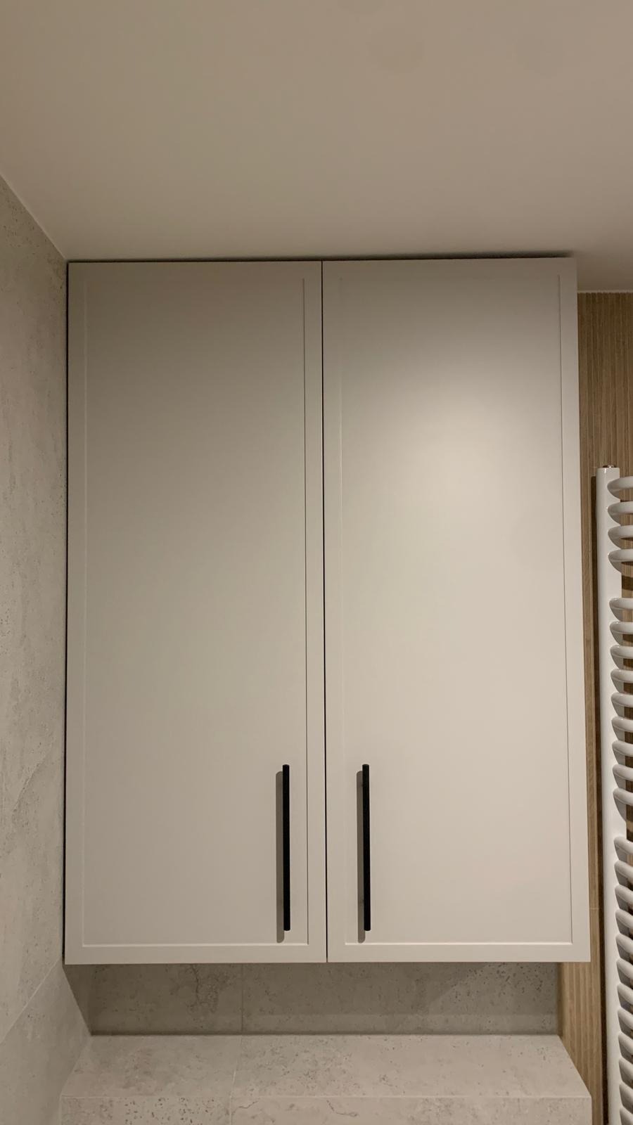 Białe szafki łazienkowe podwieszane z długimi czarnymi uchwytami, zamontowane nad blatem z szarego kamienia.