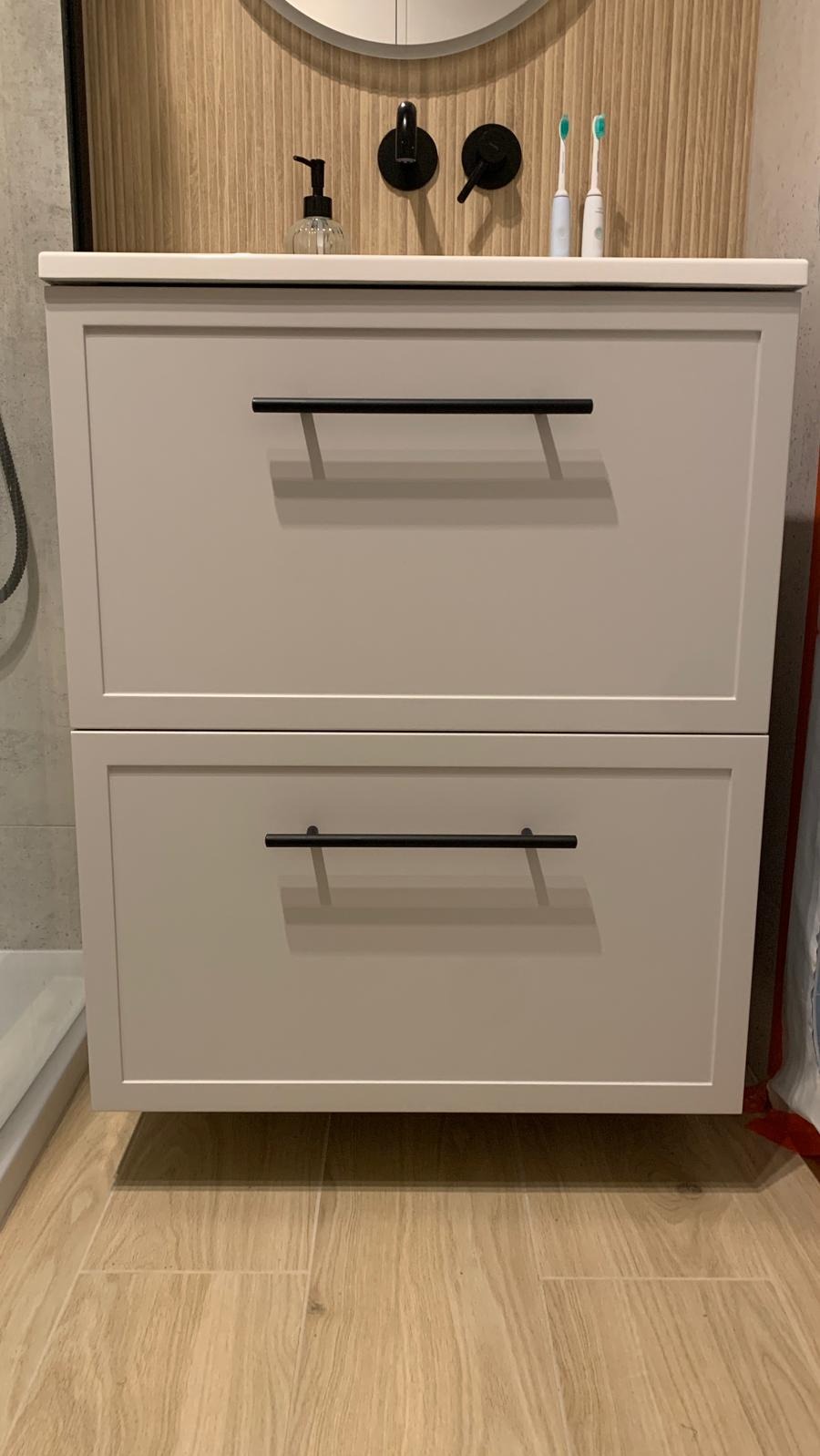 Szara szafka łazienkowa z dwiema szufladami i czarnymi, poziomymi uchwytami, stojąca na drewnopodobnych płytkach, z czarną armaturą i akcesoriami na blacie.