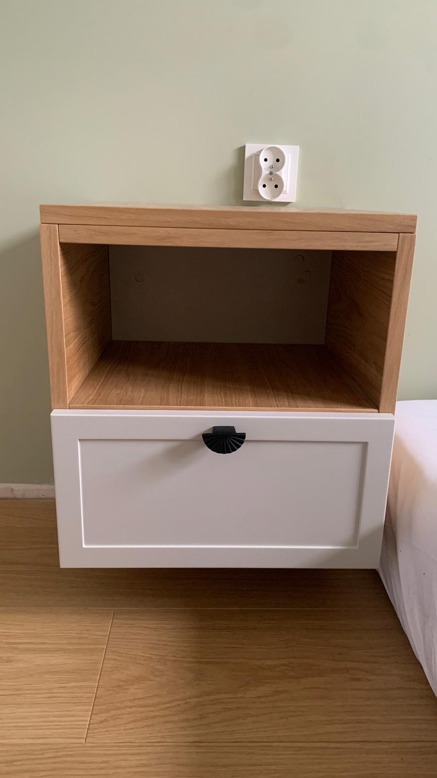 Półka nocna z szufladą, połączenie drewna w naturalnym odcieniu i białego frontu, czarny, półokrągły uchwyt, widoczne gniazdko elektryczne na ścianie.