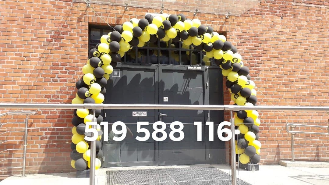 Wejście do budynku ozdobione łukiem z czarno-żółtych balonów z logo firmy, numer telefonu na pierwszym planie, ceglana ściana w tle.