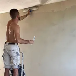 Mężczyzna bez koszulki na drabinie nakłada gładź na ścianę za pomocą szpachelki i pacy, widoczny fragment sufitu i podłogi w trakcie prac.