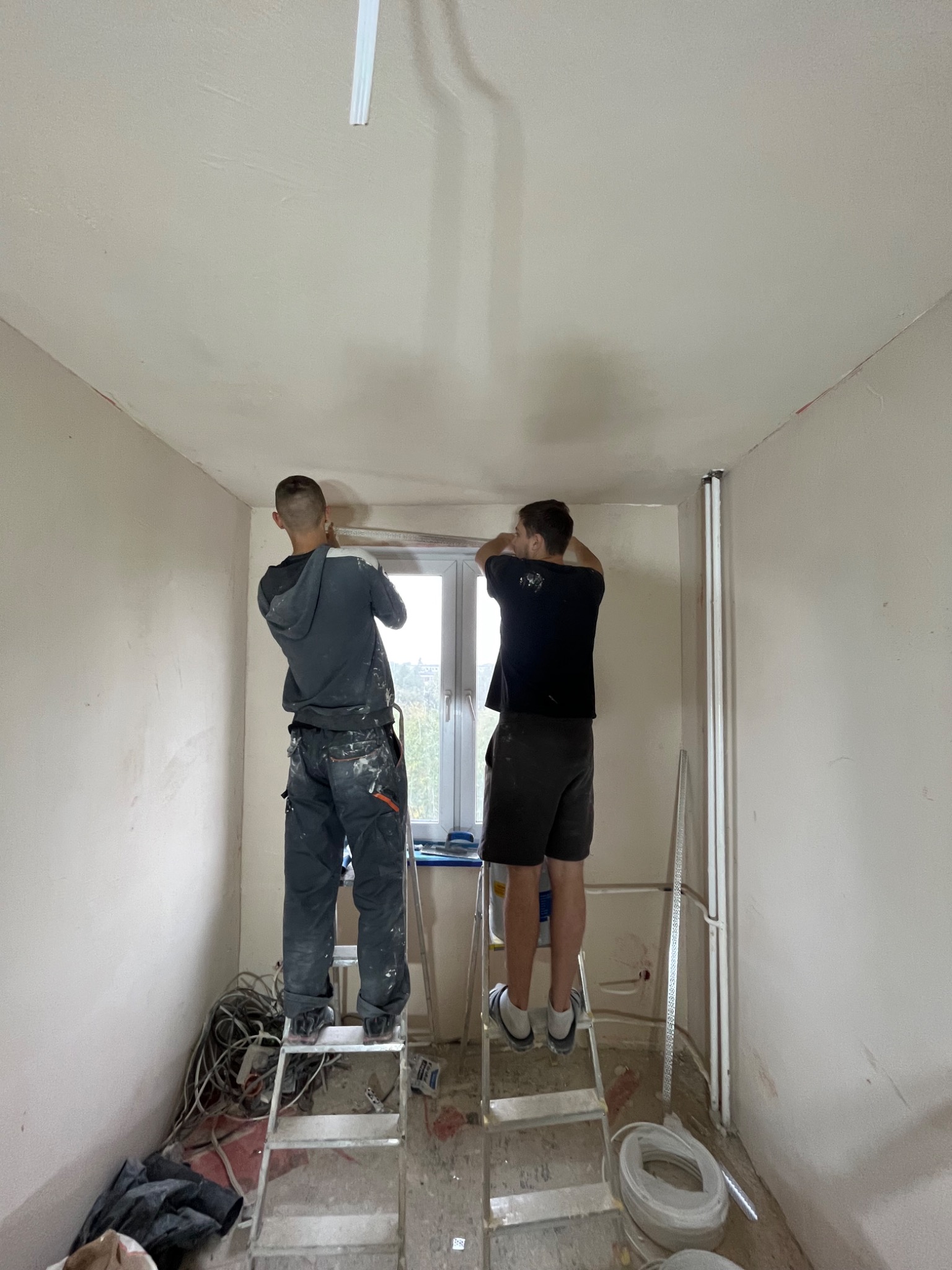 Dwóch mężczyzn na drabinach wyrównuje ścianę w pokoju podczas prac wykończeniowych, widoczne przewody elektryczne i narzędzia.