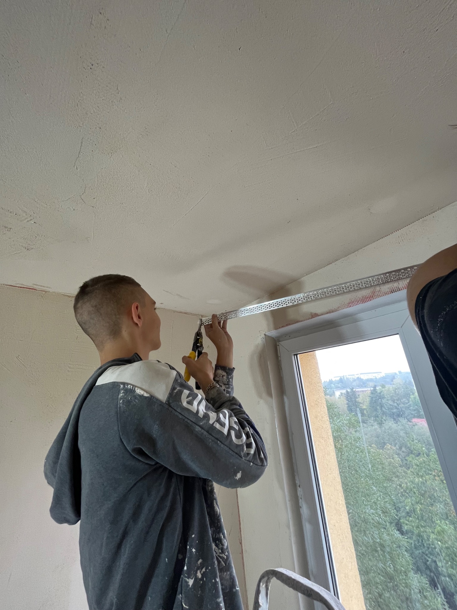 Młody mężczyzna w bluzie z plamami farby przycina nożycami metalowy profil na suficie w pobliżu okna podczas prac wykończeniowych.