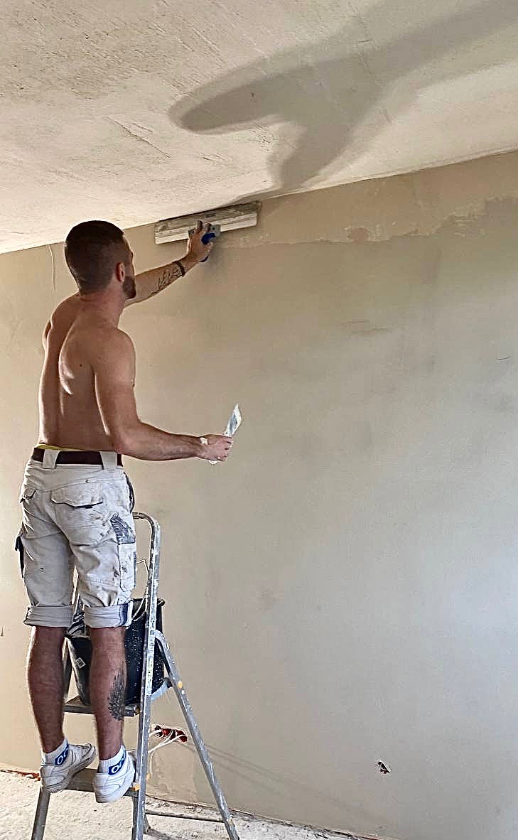 Mężczyzna bez koszulki na drabinie nakłada gładź na ścianę za pomocą szpachelki i pacy, widoczny fragment sufitu i podłogi w trakcie prac.