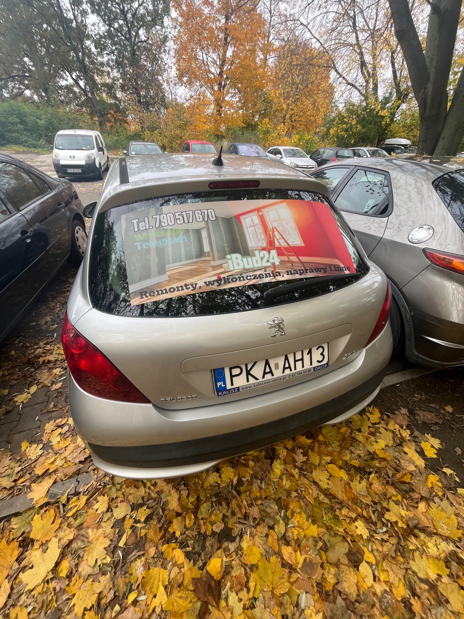 Srebrny samochód Peugeot zaparkowany na parkingu, na tylnej szybie naklejka reklamowa firmy iBud24 oferującej usługi remontowe i wykończeniowe z numerem telefonu i logotypem WhatsApp i Telegram...