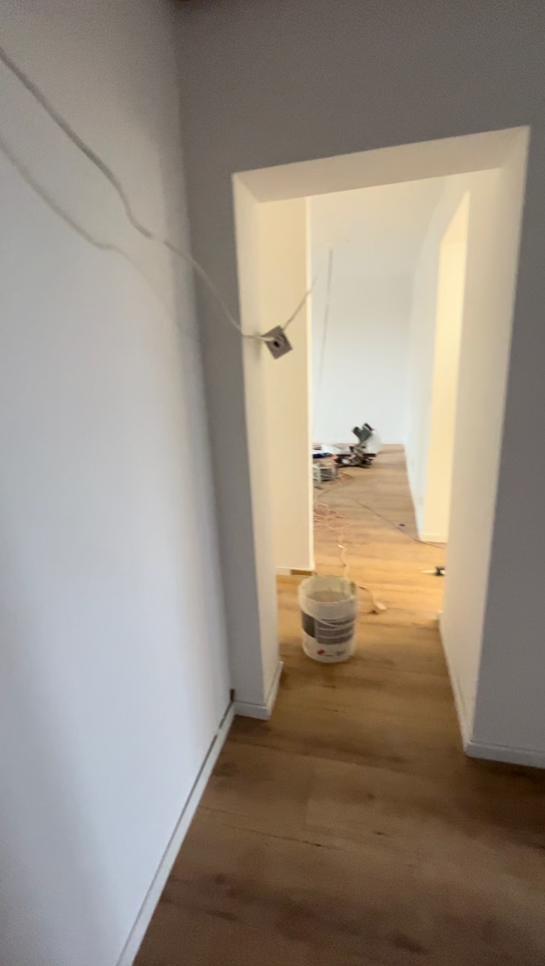 Wnętrze w trakcie prac przygotowawczych: widok przez przejście z widoczną puszką farby na podłodze, wystającymi przewodami elektrycznymi i sprzętem w tle.