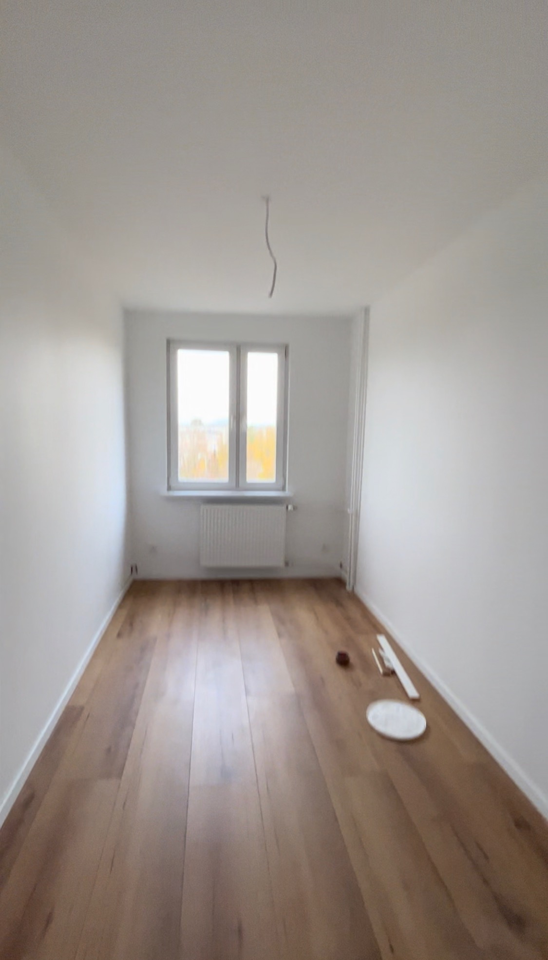 Pusty pokój po remoncie z nową drewnianą podłogą, białymi ścianami i grzejnikiem pod oknem. Na podłodze leżą akcesoria malarskie: kuweta i listwa.