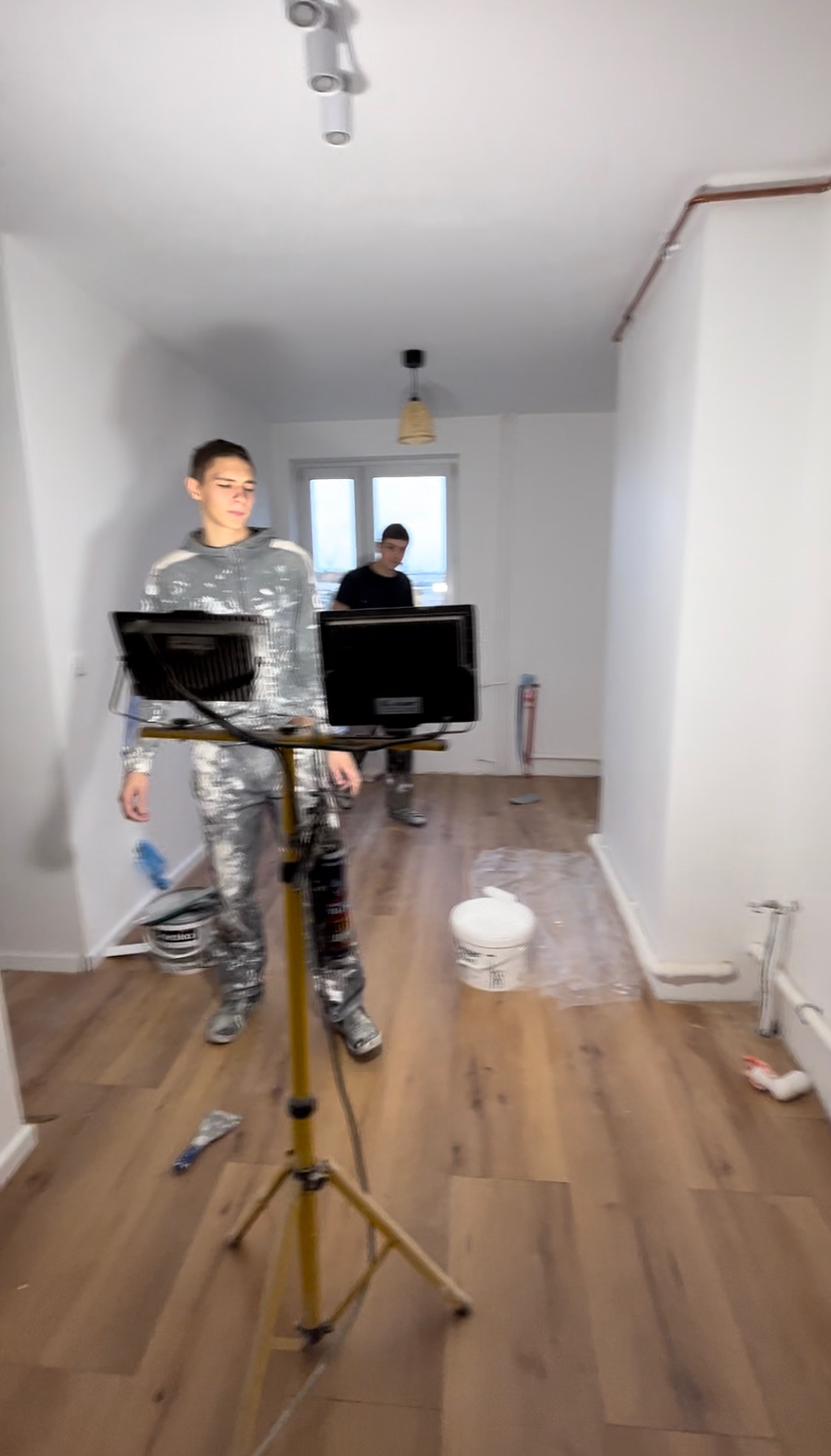 Dwóch malarzy w trakcie pracy w pustym pomieszczeniu z drewnianą podłogą, widoczny statyw z oświetleniem, wiaderka z farbą i narzędzia malarskie.