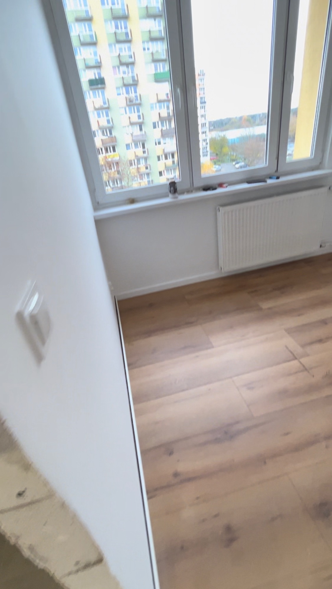 Fragment pokoju po odświeżeniu: nowa podłoga z paneli w odcieniach brązu, świeżo pomalowana biała ściana z widocznym gniazdkiem elektrycznym oraz okno z grzejnikiem i widokiem na budynek mieszkalny.