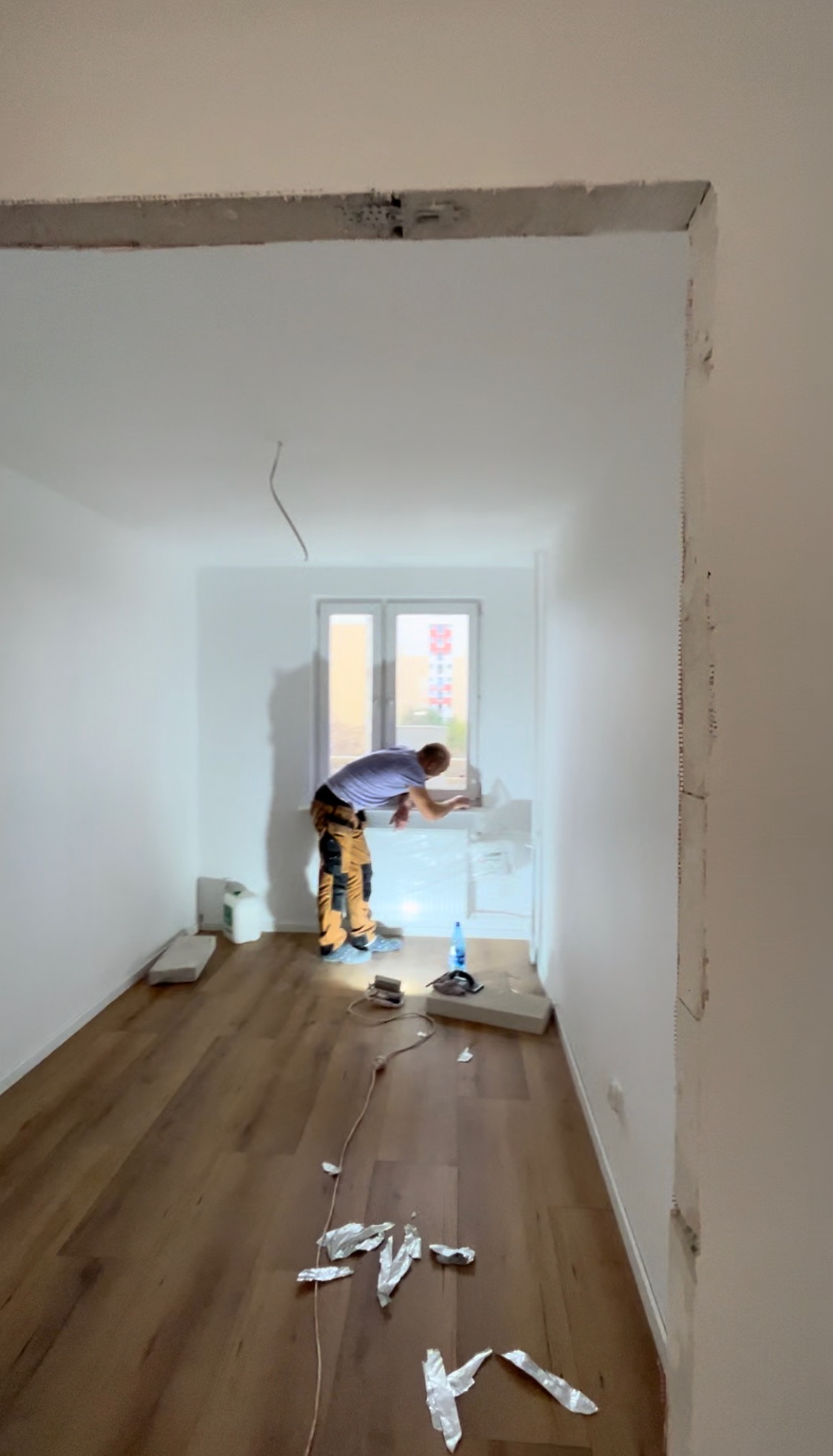 Pracownik w spodniach roboczych wygładza ścianę przy oknie w trakcie prac wykończeniowych; widoczne narzędzia, materiały i folia ochronna na podłodze z paneli.