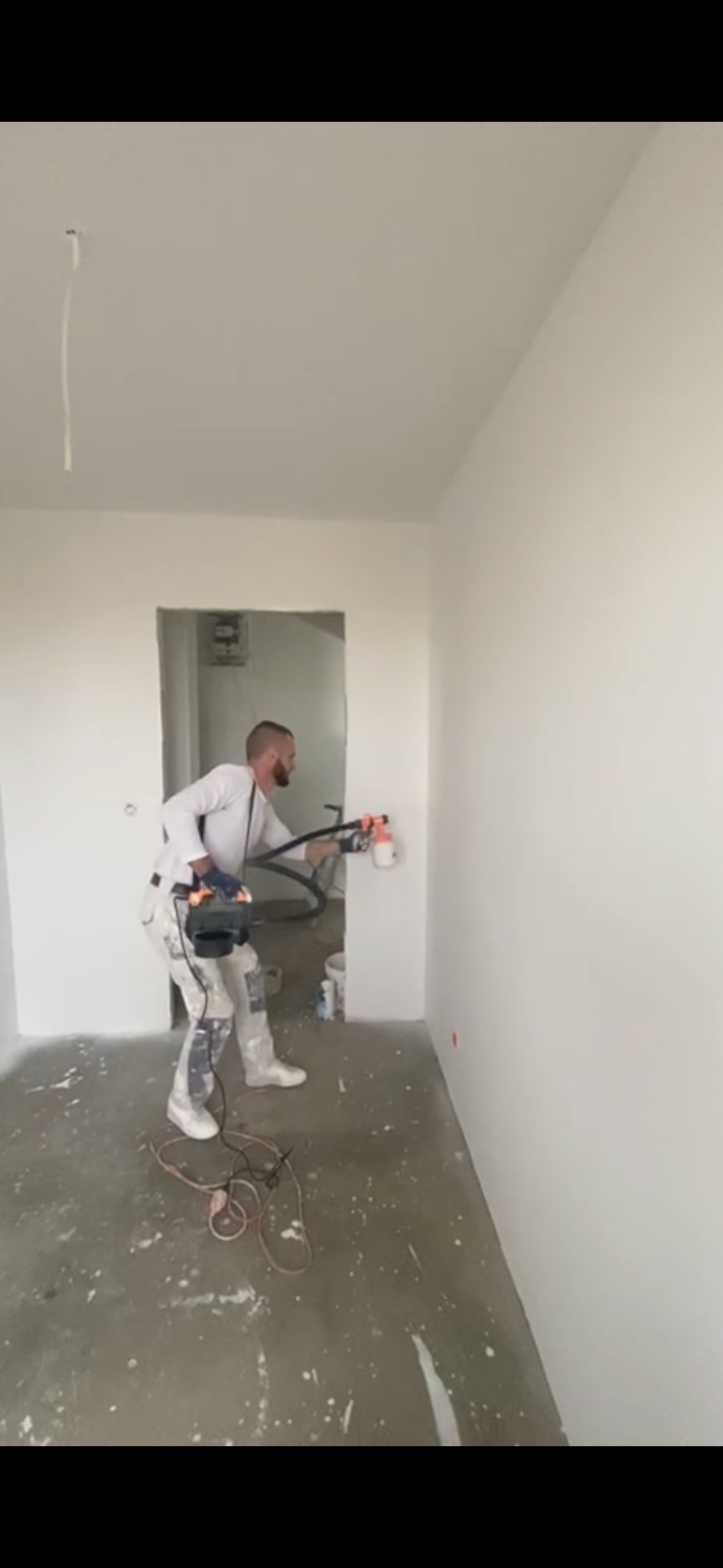 Mężczyzna w ubraniu roboczym maluje ściany na biało za pomocą elektrycznego pistoletu natryskowego w surowym pomieszczeniu z betonową podłogą.
