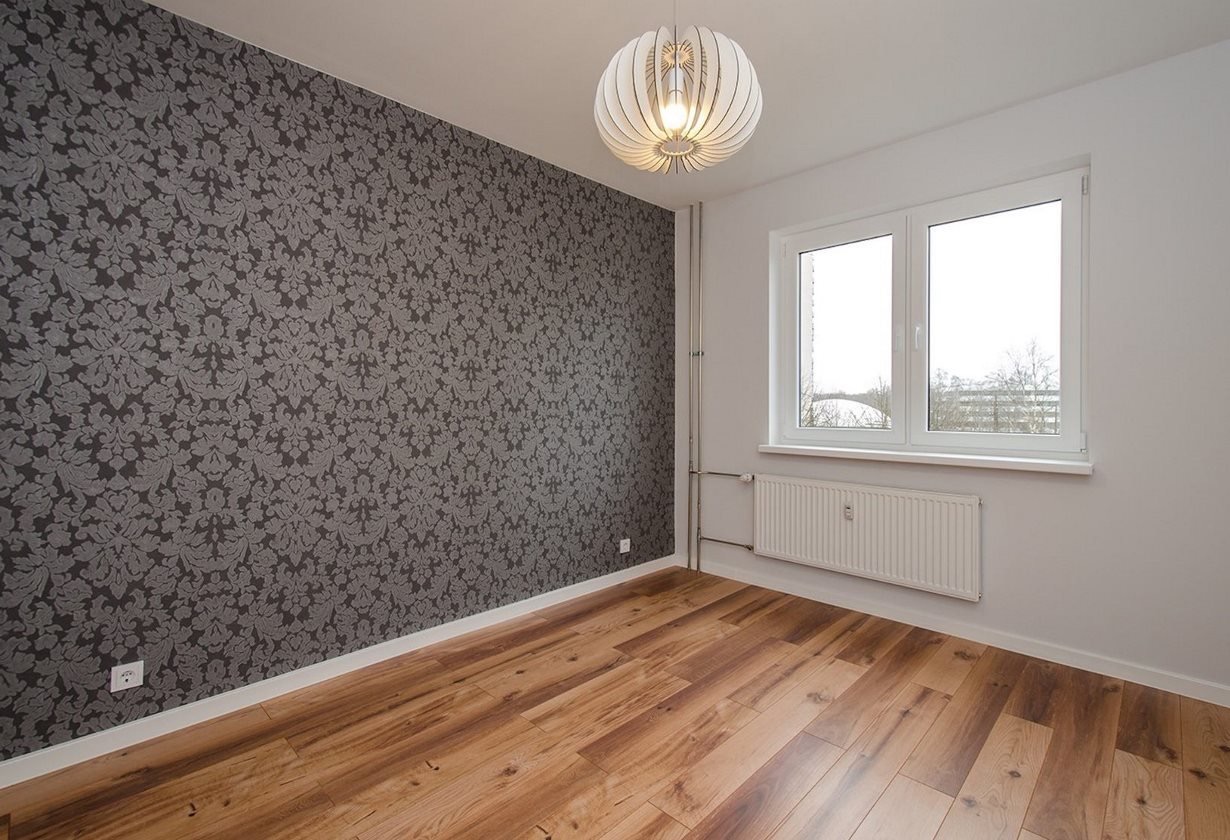 Pokój po odświeżeniu: ściana z szarą tapetą w ornamenty, drewniana podłoga, białe okno z widokiem na zewnątrz i designerska lampa wisząca.