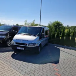 Dwa busy zaparkowane na brukowanym placu w słoneczny dzień; srebrny bus z napisem KUKULBUS.PL na przedniej szybie.