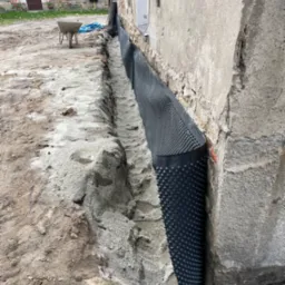 Odkryty fundament starego budynku z nałożoną czarną folią izolacyjną z wypustkami, widoczny wąski wykop i taczka w tle.