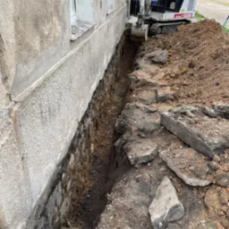 Bobcat X328 kopie wąski rów wzdłuż fundamentu starego budynku z oknami, odsłaniając ceglaną konstrukcję poniżej poziomu gruntu.