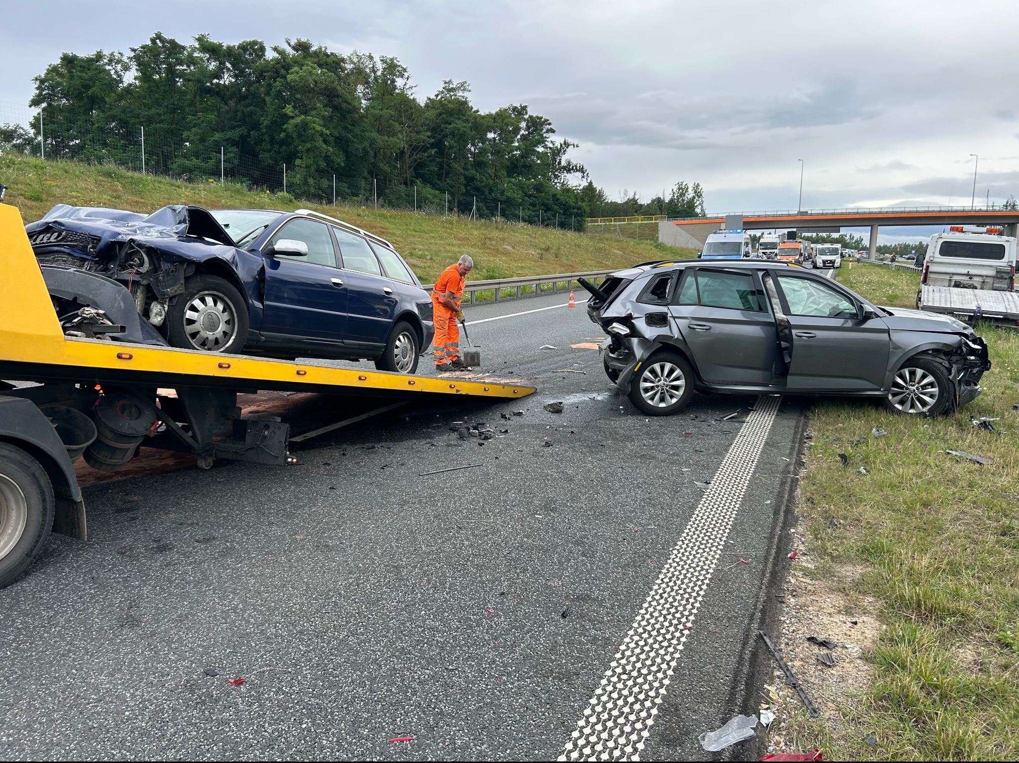 Uszkodzone samochody po wypadku na autostradzie, jeden załadowany na żółtą lawetę, pracownik w pomarańczowym kombinezonie zabezpiecza miejsce zdarzenia, widoczne odłamki i uszkodzenia karoserii.