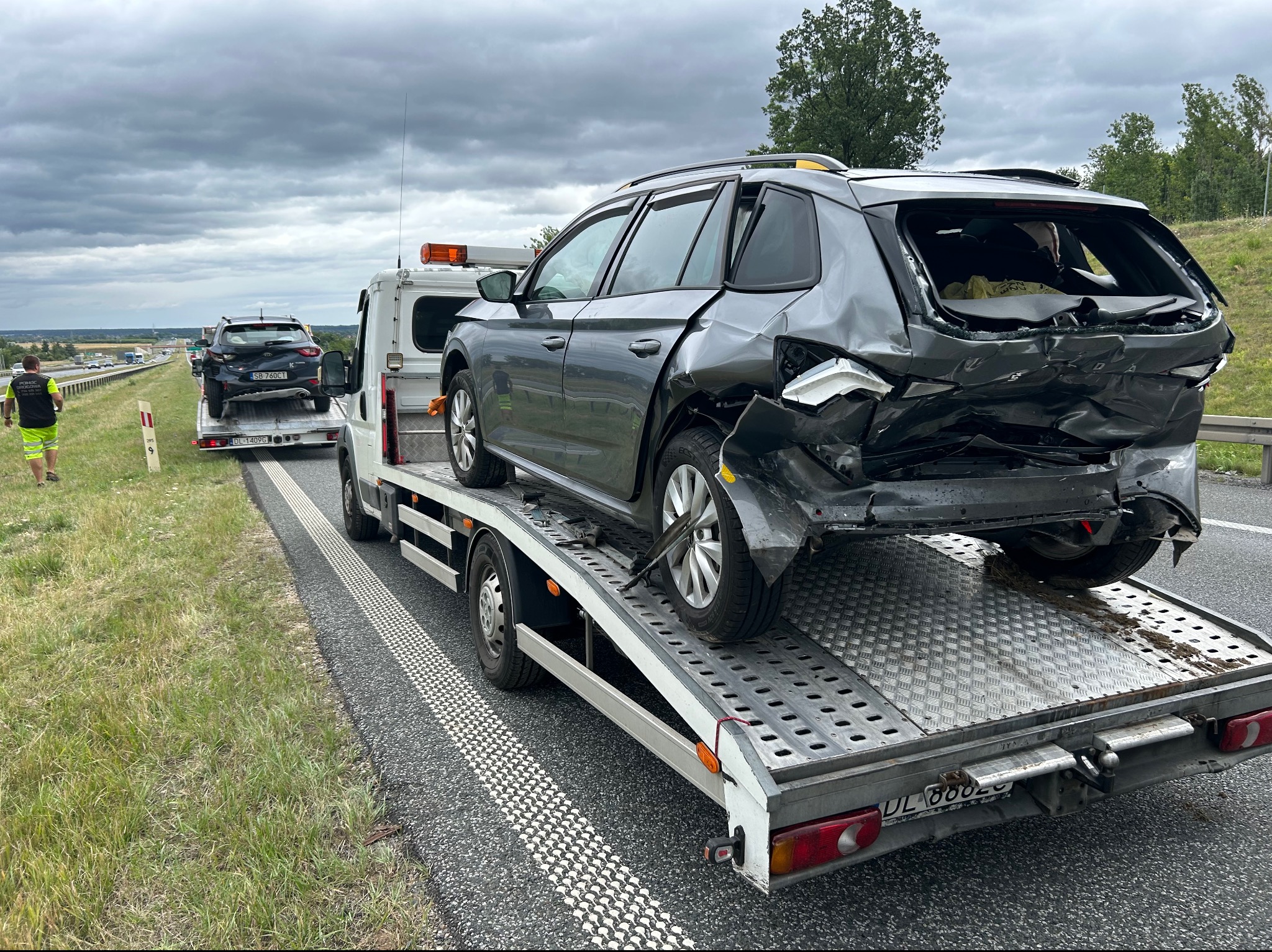 Uszkodzony szary samochód osobowy z otwartym dachem, zgniecionym tyłem i wybitymi szybami, przewożony na lawecie na autostradzie. W tle widoczny drugi pojazd na lawecie i pracownik w odblaskowej...