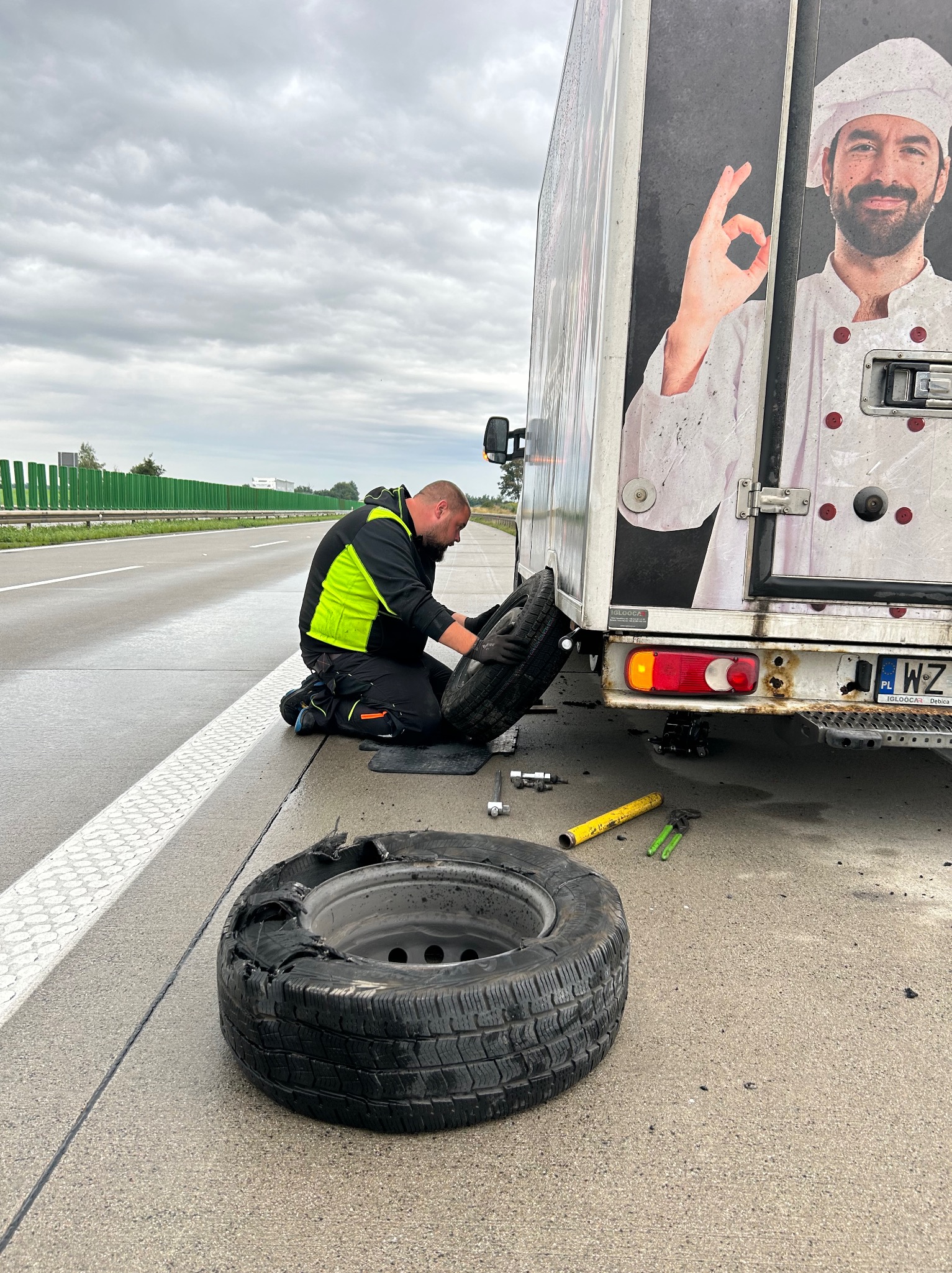 Mężczyzna w odblaskowej kurtce wymienia koło w busie na poboczu autostrady, obok leży zniszczona opona, na boku busa reklama z wizerunkiem kucharza.