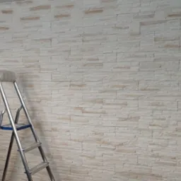 Ściana wykończona dekoracyjną płytką imitującą kamień, w odcieniach bieli i beżu, z widoczną aluminiową drabiną i rolką taśmy malarskiej na górnym szczeblu, sugerująca prace wykończeniowe.