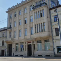 Elewacja odrestaurowanej, jasnej kamienicy z elementami sztukaterii, balkonem i wysokimi oknami, widziana z poziomu ulicy z widocznymi torami tramwajowymi.