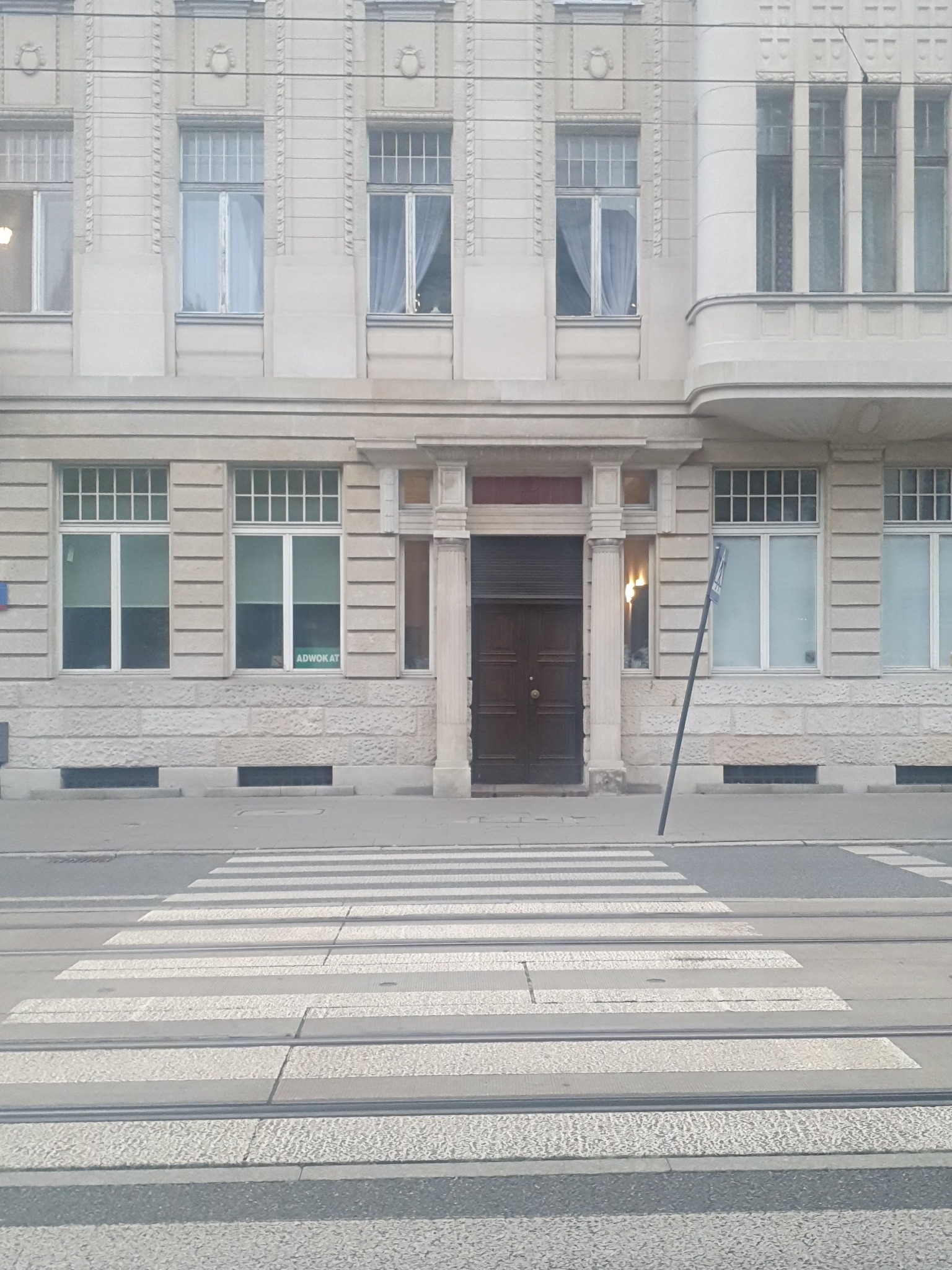 Fasada kamienicy z widocznym wejściem, oknami z firankami i napisem 'ADWOKAT' na jednej z szyb, sfotografowana z poziomu przejścia dla pieszych.