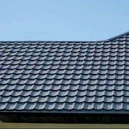 Nowy, ciemnoszary dach z metalowej dachówki modułowej, komin z blachy i okno dachowe na tle błękitnego nieba.